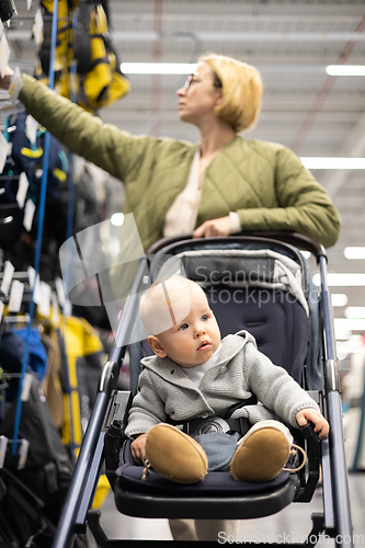 Image of Casualy dressed mother choosing sporty shoes and clothes products in sports department of supermarket store with her infant baby boy child in stroller.