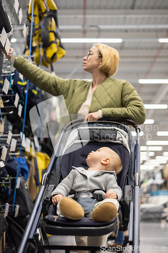 Image of Casualy dressed mother choosing sporty shoes and clothes products in sports department of supermarket store with her infant baby boy child in stroller.