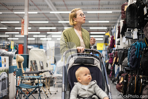 Image of Casualy dressed mother choosing sporty shoes and clothes products in sports department of supermarket store with her infant baby boy child in stroller.