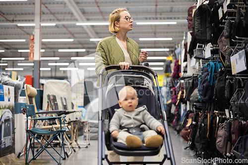 Image of Casualy dressed mother choosing sporty shoes and clothes products in sports department of supermarket store with her infant baby boy child in stroller.