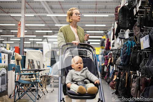 Image of Casualy dressed mother choosing sporty shoes and clothes products in sports department of supermarket store with her infant baby boy child in stroller.