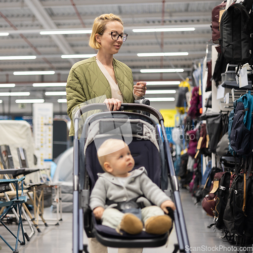 Image of Casualy dressed mother choosing sporty shoes and clothes products in sports department of supermarket store with her infant baby boy child in stroller.