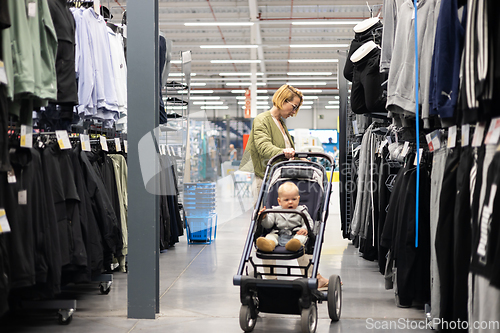 Image of Casualy dressed mother choosing sporty shoes and clothes products in sports department of supermarket store with her infant baby boy child in stroller.