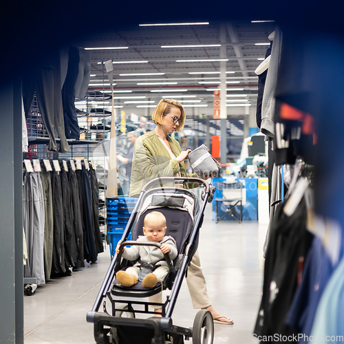 Image of Casualy dressed mother choosing sporty shoes and clothes products in sports department of supermarket store with her infant baby boy child in stroller.