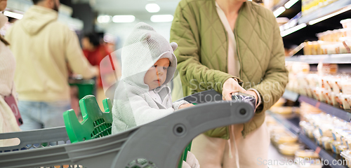 Image of Casualy dressed mother choosing products in department of supermarket grocery store with her infant baby boy child in shopping cart.