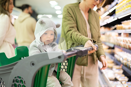 Image of Casualy dressed mother choosing products in department of supermarket grocery store with her infant baby boy child in shopping cart.