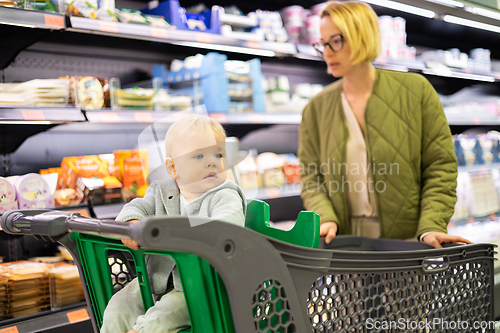 Image of Casualy dressed mother choosing canned products in department of supermarket grocery store with her infant baby boy child in shoping cart