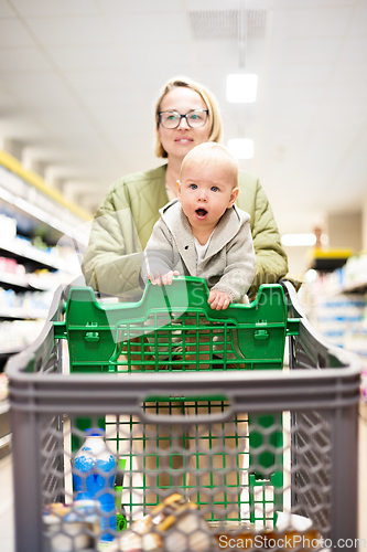 Image of Mother pushing shopping cart with her infant baby boy child down department aisle in supermarket grocery store. Shopping with kids concept.