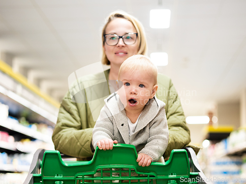 Image of Mother pushing shopping cart with her infant baby boy child down department aisle in supermarket grocery store. Shopping with kids concept.