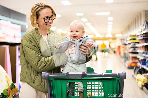 Image of Mother pushing shopping cart with her infant baby boy child down department aisle in supermarket grocery store. Shopping with kids concept.