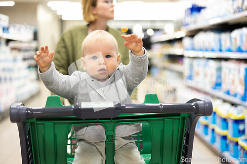 Image of Mother pushing shopping cart with her infant baby boy child down department aisle in supermarket grocery store. Shopping with kids concept.