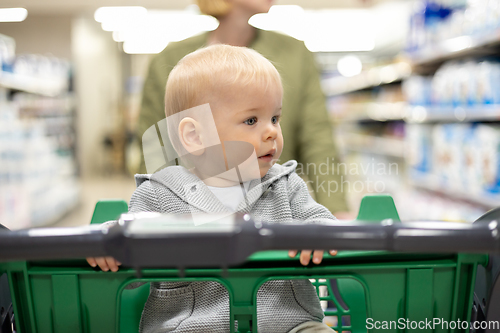 Image of Mother pushing shopping cart with her infant baby boy child down department aisle in supermarket grocery store. Shopping with kids concept.