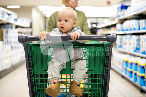 Image of Mother pushing shopping cart with her infant baby boy child down department aisle in supermarket grocery store. Shopping with kids concept.