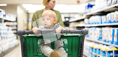 Image of Mother pushing shopping cart with her infant baby boy child down department aisle in supermarket grocery store. Shopping with kids concept.