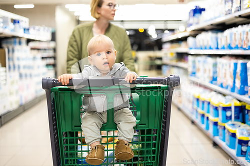 Image of Mother pushing shopping cart with her infant baby boy child down department aisle in supermarket grocery store. Shopping with kids concept.