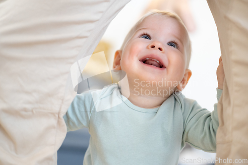 Image of Portrait of adorable cheerful infant baby boy child taking first steps holding to father's pants at home. Cute baby boy learning to walk.
