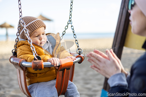 Image of Mother pushing her infant baby boy child on a swing on sandy beach playground outdoors on nice sunny cold winter day in Malaga, Spain.