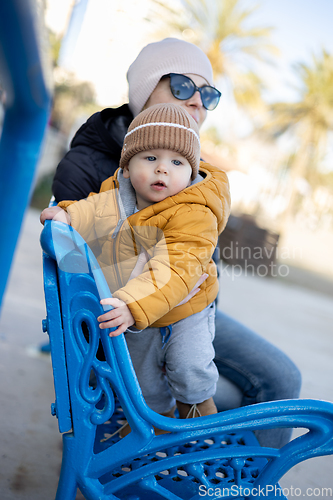 Image of Young mother with her cute infant baby boy child on bench in city park.