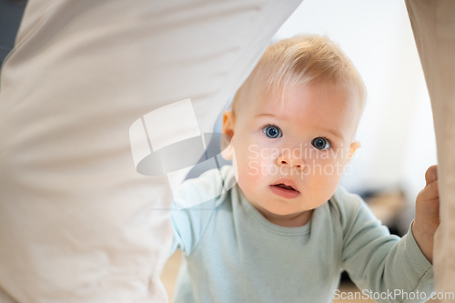 Image of Portrait of adorable curious infant baby boy child taking first steps holding to father's pants at home. Cute baby boy learning to walk.