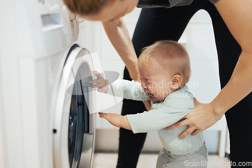 Image of Little infant baby boy child demanding mother's attention while she is multitasking, trying to do some household chores in kitchen at home. Mother on maternity leave.