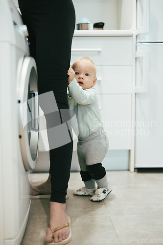 Image of Little infant baby boy child hiding between mothers legs demanding her attention while she is multitasking, trying to do some household chores in kitchen at home. Mother on maternity leave.