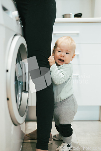 Image of Little infant baby boy child hiding between mothers legs demanding her attention while she is multitasking, trying to do some household chores in kitchen at home. Mother on maternity leave.