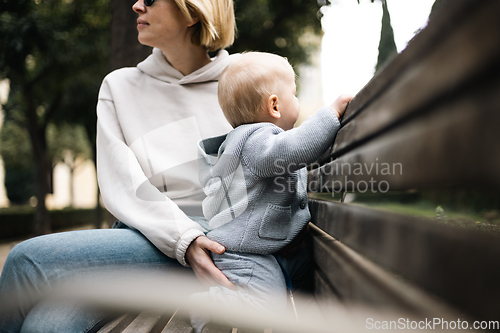 Image of Young mother with her cute infant baby boy child sitting on bench in city park.