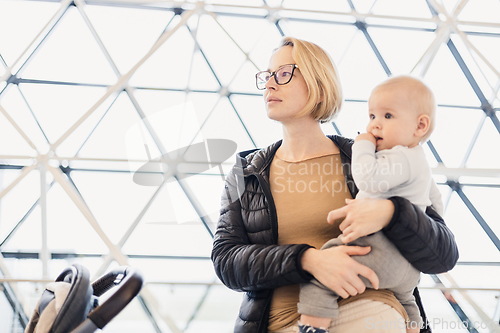 Image of Mother carying his infant baby boy child, pushing stroller at airport departure terminal waiting at boarding gates to board an airplane. Family travel with baby concept.