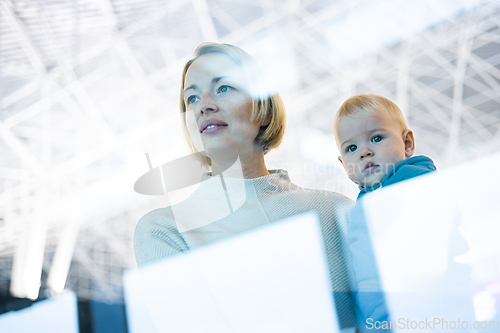 Image of Thoughtful young mother looking trough window holding his infant baby boy child while waiting to board an airplane at airport terminal departure gates. Travel with baby concept.