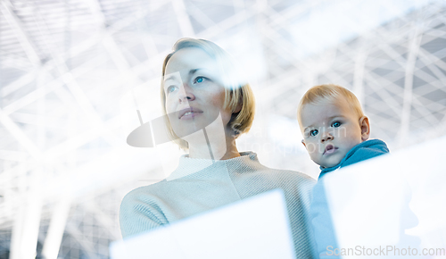 Image of Thoughtful young mother looking trough window holding his infant baby boy child while waiting to board an airplane at airport terminal departure gates. Travel with baby concept.