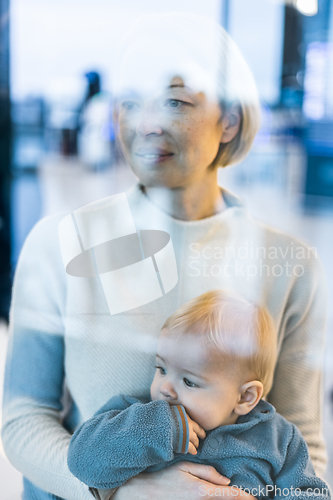 Image of Thoughtful young mother looking trough window holding his infant baby boy child while waiting to board an airplane at airport terminal departure gates. Travel with baby concept.