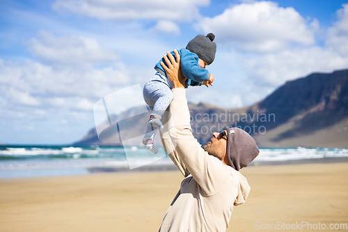 Image of Father enjoying pure nature holding and playing with his infant baby boy son in on windy sandy beach of Famara, Lanzarote island, Spain. Family travel and parenting concept.