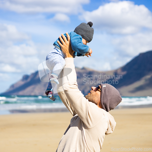 Image of Father enjoying pure nature holding and playing with his infant baby boy son in on windy sandy beach of Famara, Lanzarote island, Spain. Family travel and parenting concept.