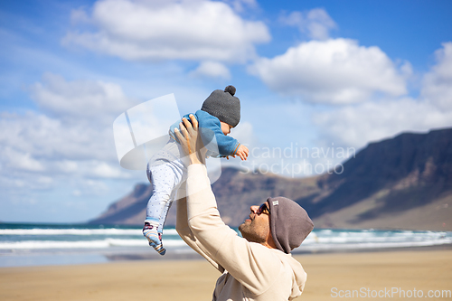 Image of Father enjoying pure nature holding and playing with his infant baby boy son in on windy sandy beach of Famara, Lanzarote island, Spain. Family travel and parenting concept.