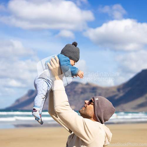 Image of Father enjoying pure nature holding and playing with his infant baby boy son in on windy sandy beach of Famara, Lanzarote island, Spain. Family travel and parenting concept.