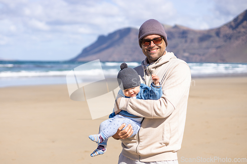 Image of Father enjoying pure nature holding and playing with his infant baby boy son in on windy sandy beach of Famara, Lanzarote island, Spain. Family travel and parenting concept.