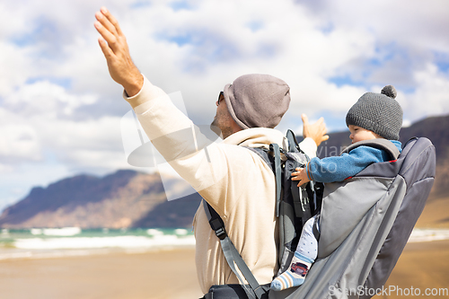 Image of Young father rising hands to the sky while enjoying pure nature carrying his infant baby boy son in backpack on windy sandy beach. Family travel concept.