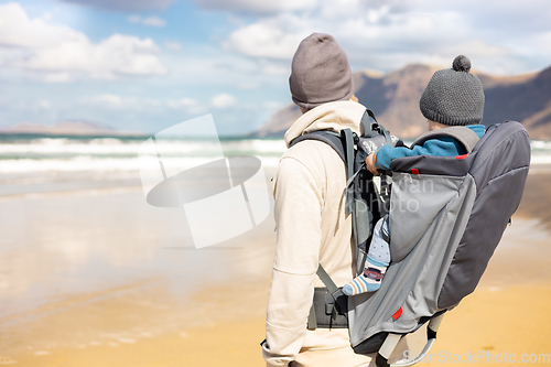 Image of Young father carrying his infant baby boy son in backpack on windy sandy beach. Family travel and winter vacation concept.