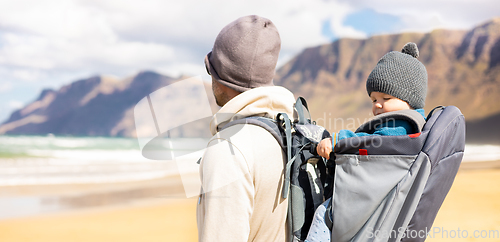 Image of Young father carrying his infant baby boy in backpack on windy sandy beach. Family travel and vacation concept.