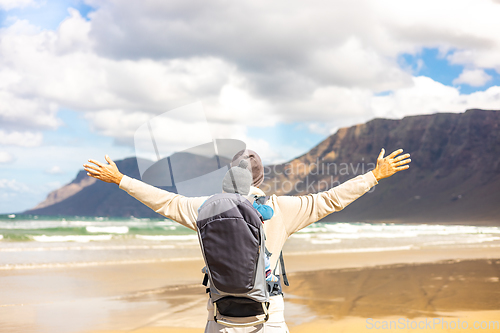 Image of Young father rising hands to the sky while enjoying pure nature carrying his infant baby boy son in backpack on windy sandy beach of Famara, Lanzarote island, Spain. Family travel concept.