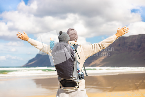 Image of Young father rising hands to the sky while enjoying pure nature carrying his infant baby boy son in backpack on windy sandy beach of Famara, Lanzarote island, Spain. Family travel concept.