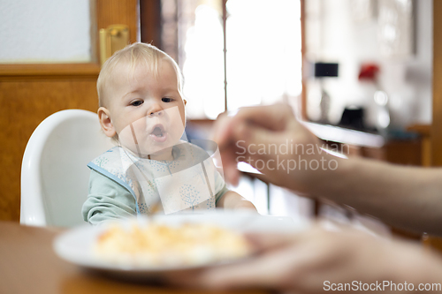 Image of Mother spoon feeding her infant baby boy child sitting in high chair at the dining table in kitchen at home