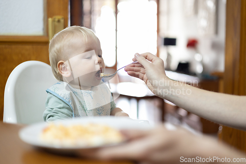 Image of Mother spoon feeding her infant baby boy child sitting in high chair at the dining table in kitchen at home