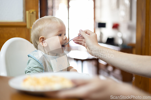 Image of Mother spoon feeding her infant baby boy child sitting in high chair at the dining table in kitchen at home