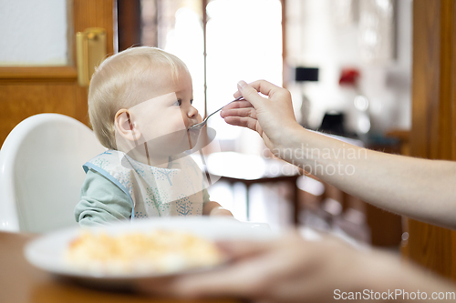 Image of Mother spoon feeding her infant baby boy child sitting in high chair at the dining table in kitchen at home
