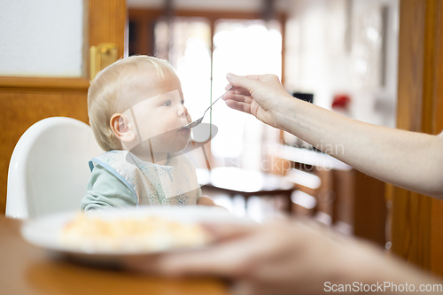 Image of Mother spoon feeding her infant baby boy child sitting in high chair at the dining table in kitchen at home