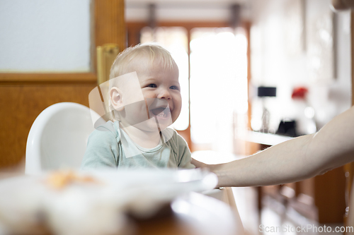 Image of Adorable cheerful happy infant baby boy child smiling while sitting in high chair at the dining table in kitchen at home beeing spoon fed by his mother