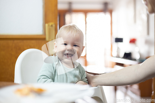 Image of Adorable cheerful happy infant baby boy child smiling while sitting in high chair at the dining table in kitchen at home beeing spoon fed by his mother.