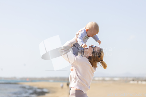 Image of Mother enjoying summer vacations holding, playing and lifting his infant baby boy son high in the air on sandy beach on Lanzarote island, Spain. Family travel and vacations concept.