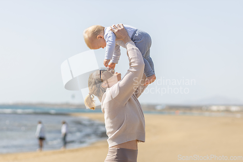 Image of Mother enjoying summer vacations holding, playing and lifting his infant baby boy son high in the air on sandy beach on Lanzarote island, Spain. Family travel and vacations concept.